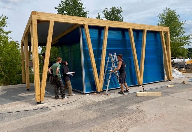 Eine Gruppe von vier Personen steht neben einem halbfertigen Gebäude mit blauen Wänden und Holzrahmen, das an ein Projekt der Hochschule Coburg erinnert. Eine Person steht auf einer Leiter, die anderen diskutieren auf dem Boden, im Hintergrund sind Bäume und Baumaterialien zu sehen.