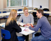 Drei Personen sitzen an einem Tisch im Hörsaal der Hochschule Coburg und diskutieren angeregt. Mit Notizbüchern und einem offenen Buch tauschen sie zwischen Tischen und Stühlen Ideen aus, im Hintergrund fällt Sonnenlicht durch große Fenster.