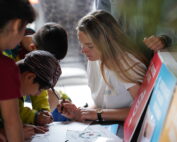 Eine Gruppe Kinder versammelt sich um eine Frau, die an der Hochschule Coburg auf Papier zeichnet. Die Szene ist lebhaft und farbenfroh, in der Nähe hängen Plakate. Die Kinder beteiligen sich eifrig und schauen zu, wodurch eine Atmosphäre des Lernens und der Kreativität entsteht.