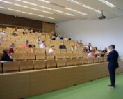 Ein Dozent steht vor einem Hörsaal der Hochschule Coburg und spricht zu einem Publikum, das in Reihen von Holzbänken sitzt. Die Teilnehmer sind über den geräumigen Raum verteilt und hören aufmerksam zu.