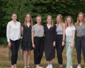 Sieben Frauen stehen in einer Reihe im Freien und lächeln in die Kamera. In lässiger, aber dennoch stilvoller Kleidung posieren sie stolz vor einer Kulisse aus grünen Bäumen und Gras und repräsentieren die Hochschule Coburg mit Anmut und Kameradschaft.