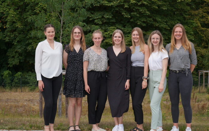 Sieben Frauen stehen in einer Reihe im Freien und lächeln in die Kamera. In lässiger, aber dennoch stilvoller Kleidung posieren sie stolz vor einer Kulisse aus grünen Bäumen und Gras und repräsentieren die Hochschule Coburg mit Anmut und Kameradschaft.