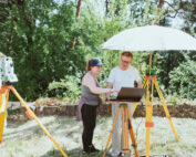 Zwei Personen arbeiten draußen an der Hochschule Coburg mit Vermessungsgeräten. Einer verwendet einen Laptop auf einem Stativ mit Sonnenschutz, während der andere auf den Bildschirm zeigt. In der Nähe steht ein weiteres Vermessungsgerät, Bäume bilden eine ruhige Kulisse.