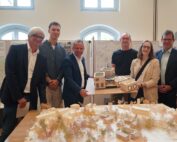 Sieben Menschen stehen stolz zusammen und halten Architekturmodelle in einem Raum der Hochschule Coburg. Große Fenster beleuchten die im Hintergrund auf Tafeln hängenden Poster und schaffen so eine inspirierende Atmosphäre für Kreativität und Zusammenarbeit.