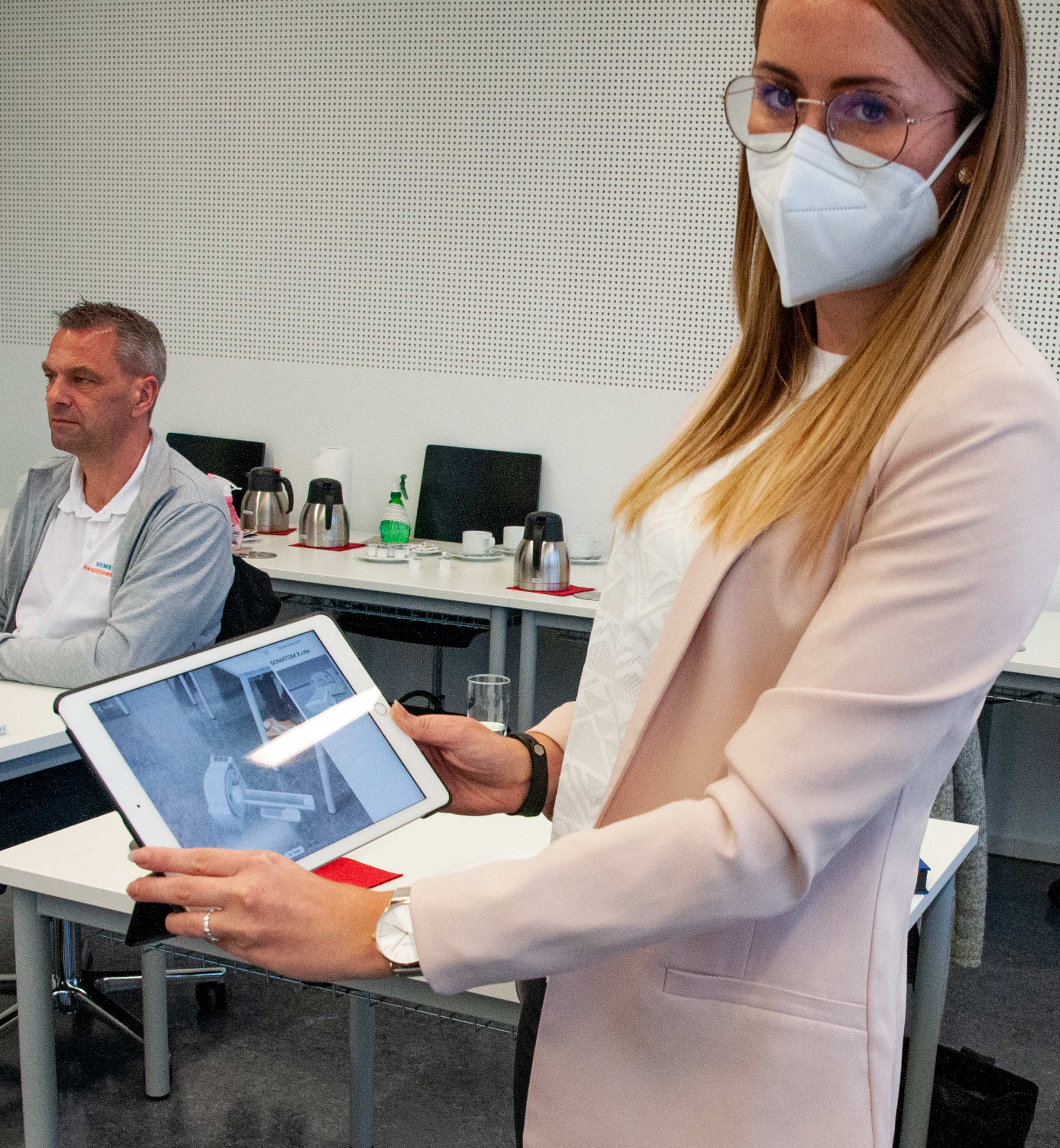 Eine Frau mit Maske und Brille hält ein Tablet mit einem Schwarz-Weiß-Bild in der Hand. Sie steht neben einer sitzenden Person in einem Hörsaal der Hochschule Coburg. Im Hintergrund sind Tische, Stühle und Kaffeeautomaten zu sehen.