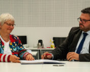 Eine Frau und ein Mann sitzen an einem Tisch und führen ein formelles Gespräch. Die Frau hält einen Stift neben einem Dokument der Hochschule Coburg, während der Mann, der eine Brille und eine Krawatte trägt, aufmerksam zuhört. Der Hintergrund ist eine schlichte, helle Wand, auf der eine Kaffeekanne zu sehen ist.