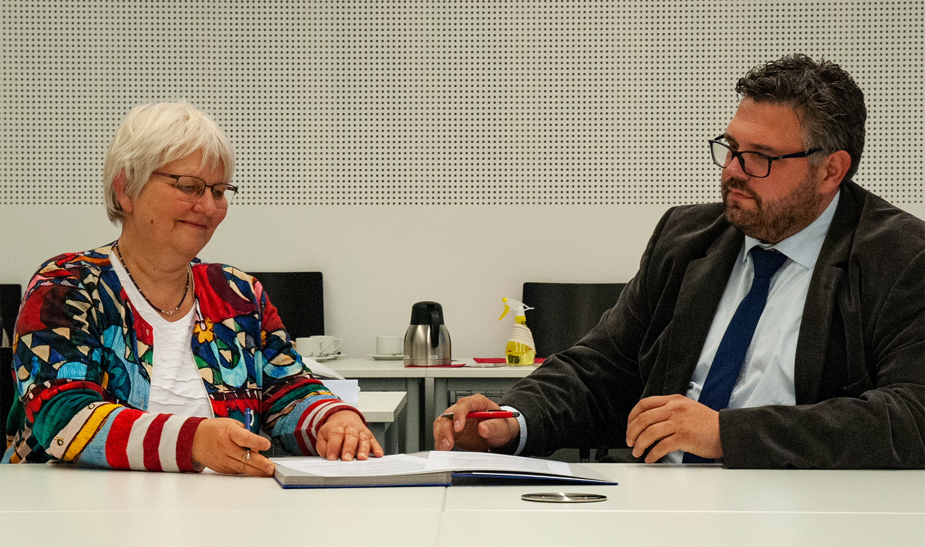Eine Frau und ein Mann sitzen an einem Tisch und führen ein formelles Gespräch. Die Frau hält einen Stift neben einem Dokument der Hochschule Coburg, während der Mann, der eine Brille und eine Krawatte trägt, aufmerksam zuhört. Der Hintergrund ist eine schlichte, helle Wand, auf der eine Kaffeekanne zu sehen ist.