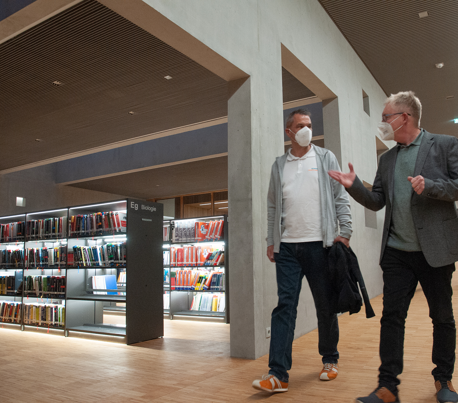 Zwei maskierte Männer gehen durch eine moderne Bibliothek der Hochschule Coburg und unterhalten sich. Sie kommen an Regalen voller Bücher über Biologie vorbei, ihre Schritte sind leise auf dem Holzboden unter der von Lichtern durchzogenen Decke.