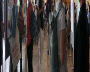 Menschen haben sich in einem Konferenzsaal der Hochschule Coburg versammelt. An Ständen hängen Poster. Die Teilnehmer diskutieren, halten Getränke in der Hand und tragen Namensschilder. Große Fenster auf einer Seite lassen Tageslicht herein, während im Hintergrund eine digitale Präsentation zu sehen ist.