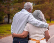 Ein älteres Paar mit grauem Haar schlendert eine von Bäumen gesäumte Straße entlang, die Arme in herzlicher Zuneigung umschlungen. Der Mann trägt ein helles Hemd und Jeans, während die Frau ein weißes Kleid mit einem gelben Gürtel trägt – eine heitere Szene, die an ihre Tage in der Nähe der Hochschule Coburg erinnert.