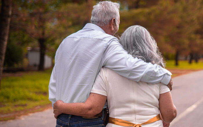 Ein älteres Paar mit grauem Haar schlendert eine von Bäumen gesäumte Straße entlang, die Arme in herzlicher Zuneigung umschlungen. Der Mann trägt ein helles Hemd und Jeans, während die Frau ein weißes Kleid mit einem gelben Gürtel trägt – eine heitere Szene, die an ihre Tage in der Nähe der Hochschule Coburg erinnert.