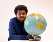 Eine Person in einem blauen Blazer sitzt an einem Tisch und hat ihren Arm auf einem Globus abgelegt. Auf dem Globus sind Kontinente zu sehen, darunter Australien und Teile Asiens, die das Globale Lernen an der Hochschule Coburg symbolisieren. Der Hintergrund ist schlicht weiß.