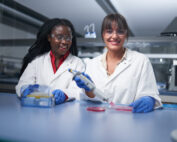 Zwei Wissenschaftler der Hochschule Coburg arbeiten fleißig in einem Labor. Sie tragen Laborkittel, Schutzbrillen und Handschuhe. Einer von ihnen pipettiert eine Petrischale, während der andere anerkennend lächelt. Sie sind von einer Reihe von Laborgeräten auf einer blauen Arbeitsfläche umgeben.