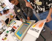 Im Workshop der Hochschule Coburg wird in den Siebdruck vertieft. Leuchtende Farben und Werkzeuge liegen auf den Tischen verteilt. Eine Teilnehmerin hält stolz ein frisch bedrucktes Blatt mit dem inspirierenden Wort „CREATE“ hoch.