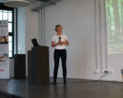 Eine Person steht auf einer Bühne der Hochschule Coburg, hält ein Mikrofon und hält eine Präsentation. Sie trägt ein weißes Hemd und eine schwarze Hose, daneben steht ein Laptop auf einem Podium. Ein projiziertes Bild eines Waldes bereichert den Hintergrund und schafft eine spannende Atmosphäre für das Publikum.