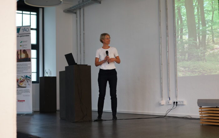 Eine Person steht auf einer Bühne der Hochschule Coburg, hält ein Mikrofon und hält eine Präsentation. Sie trägt ein weißes Hemd und eine schwarze Hose, daneben steht ein Laptop auf einem Podium. Ein projiziertes Bild eines Waldes bereichert den Hintergrund und schafft eine spannende Atmosphäre für das Publikum.