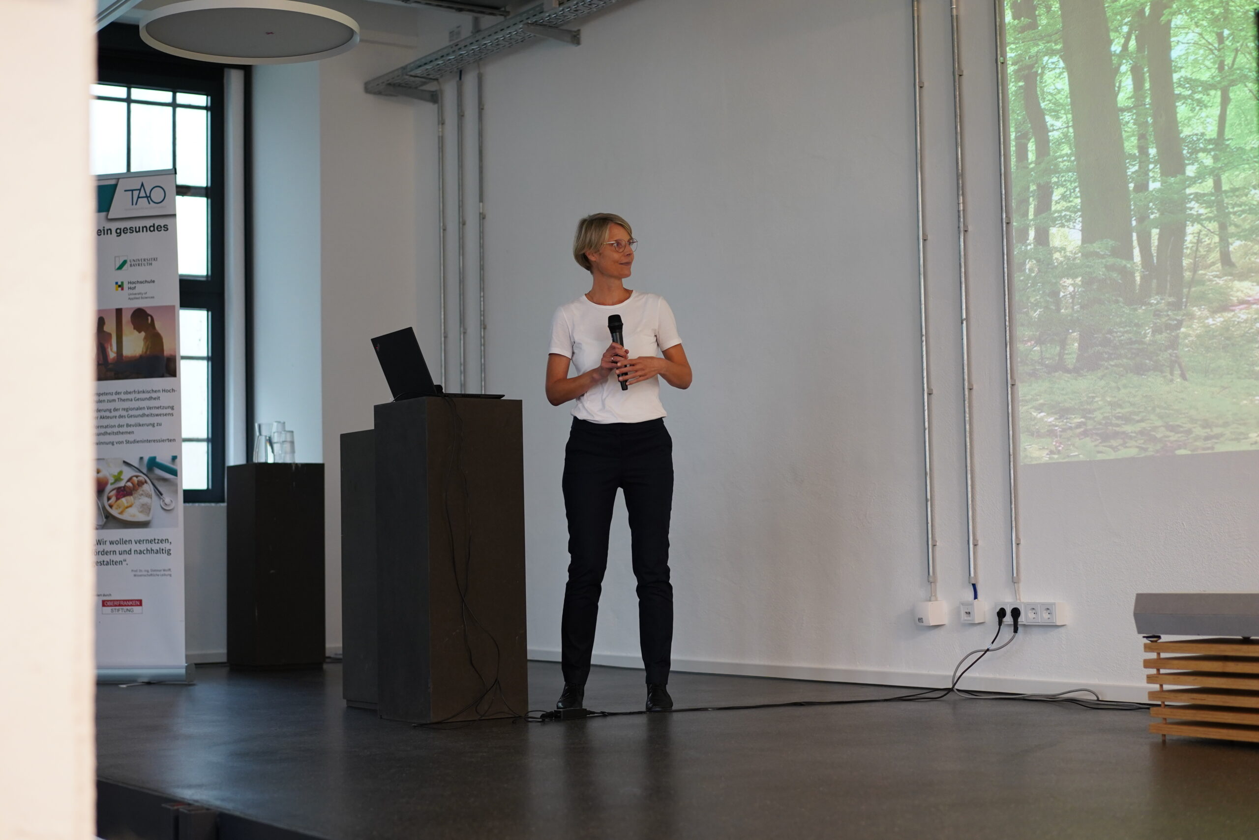 Eine Person steht auf einer Bühne der Hochschule Coburg, hält ein Mikrofon und hält eine Präsentation. Sie trägt ein weißes Hemd und eine schwarze Hose, daneben steht ein Laptop auf einem Podium. Ein projiziertes Bild eines Waldes bereichert den Hintergrund und schafft eine spannende Atmosphäre für das Publikum.