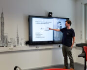 Ein Mann steht in einem Hörsaal der Hochschule Coburg und zeigt auf einen großen Monitor, auf dem eine Gleichung zur Lichtbeugung angezeigt wird. Der Raum ist mit einem Stadtbild an der Wand und modernen Möbeln ausgestattet, darunter einem roten Stuhl und einem langen Tisch.