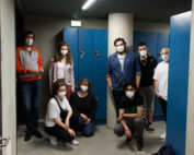 Eine Gruppe von acht maskierten Menschen steht in einem Flur der Hochschule Coburg, im Hintergrund sind blaue Schließfächer zu sehen. Einige sind leger gekleidet, andere knien, eingerahmt von Betonwänden und einer dunklen Decke.