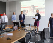 Eine Gruppe von sechs Personen steht in einem Konferenzraum der Hochschule Coburg. Sie sitzen mit etwas Abstand zueinander und lächeln, während hinter ihnen auf einer Projektionsfläche eine Website angezeigt wird. Auf dem Holztisch davor stehen mehrere Stühle und Computer.