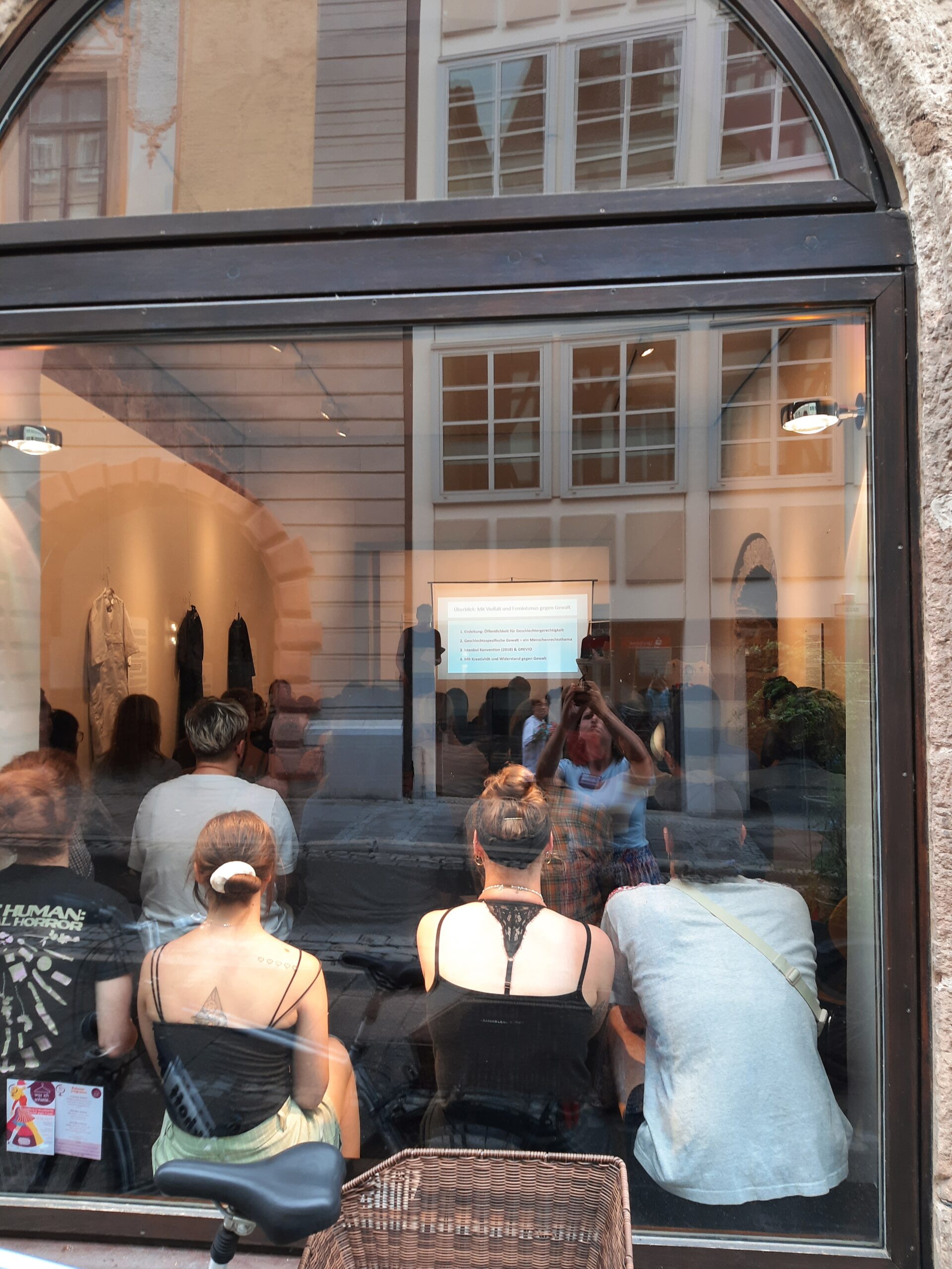 Menschen sitzen in einer Galerie oder einem Geschäft und haben den Blick durch ein großes Fenster frei. Ihr Blick geht auf einen Bildschirm, auf dem eine Präsentation der Hochschule Coburg läuft. An den Wänden um sie herum hängen Kleidungsstücke und im Glas sind Spiegelungen von Gebäuden zu sehen.