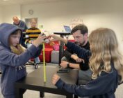 Zwei junge Mädchen messen an einem Tisch in einem Klassenzimmer der Hochschule Coburg ein gelbes Maßband. Ein Mann beobachtet sie, während eine Frau und eine weitere Person im Hintergrund zuschauen. Der Raum ist voller Unterrichtsmaterialien und verfügt über eine Wanduhr.
