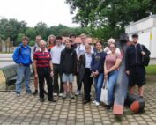 Eine Gruppe von dreizehn Personen, sowohl Männer als auch Frauen, steht entspannt draußen auf einer gepflasterten Fläche der Hochschule Coburg. Sie lächeln in die Kamera. Im Hintergrund sind Bäume und ein Gebäude zu sehen.
