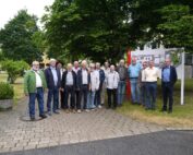 Eine Gruppe von Menschen steht draußen auf einem gepflasterten Weg an der Hochschule Coburg, im Hintergrund sind Bäume und ein Gebäude zu sehen. Sie posieren für ein Gruppenfoto, auf der rechten Seite sind etwas Grün und eine Karte oder ein Schild zu sehen.