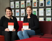 Zwei Frauen sitzen auf einem roten Sofa in der Hochschule Coburg und halten Bücher mit dem Titel „Communications Engineering Desk Reference“ in den Händen. Sie stehen vor einer Wand, die mit verschiedenen Publikationen geschmückt ist, wodurch eine akademische Atmosphäre entsteht, die an eine Bibliothek erinnert.