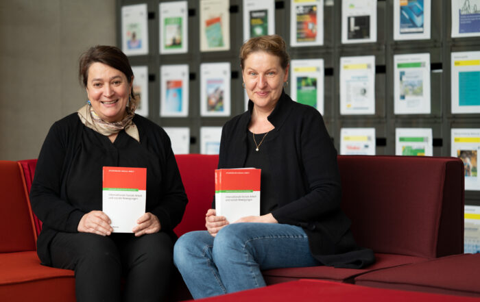 Zwei Frauen sitzen auf einem roten Sofa in der Hochschule Coburg und halten Bücher mit dem Titel „Communications Engineering Desk Reference“ in den Händen. Sie stehen vor einer Wand, die mit verschiedenen Publikationen geschmückt ist, wodurch eine akademische Atmosphäre entsteht, die an eine Bibliothek erinnert.