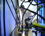 Eine Person mit Brille bedient an der Hochschule Coburg einen 3D-Drucker mit mehreren Stäben und Drähten. Im Fokus steht die Maschine, während die Person, die in dem gut beleuchteten Raum darüber steht, den komplizierten Druckvorgang scheinbar einstellt oder beobachtet.