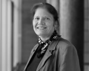 Eine Person mit langen Haaren, einem gemusterten Schal und einer Jacke steht lächelnd im Flur der Hochschule Coburg, flankiert von ihren ikonischen Säulen. Das Schwarzweißbild fängt einen Moment heiterer Eleganz ein.