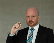 Ein bärtiger Mann in Anzug und gestreifter Krawatte, der die akademische Aura der Hochschule Coburg widerspiegelt, hält vor einem grauen Hintergrund ein kleines Fläschchen mit Flüssigkeit. Sein Gesichtsausdruck ist ernst und lässt auf die Bedeutung akademischer Forschung schließen.