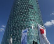 Unter dem klaren blauen Himmel der Hochschule Coburg steht ein hohes, zylindrisches Gebäude mit einem kreuzförmigen Muster aus Glas und Stahl. Im Vordergrund sind drei Flaggen zu sehen, die Logos vor einer Kulisse moderner Architektur zeigen.