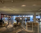 Die geräumige, moderne Lobby der Hochschule Coburg verfügt über große Fenster, die den Bereich mit natürlichem Licht durchfluten. Sie verfügt über einen eleganten Empfangstresen, Glasbarrieren und viele Menschen, die sich dort tummeln. Die dunklen Bodenfliesen bilden einen eleganten Kontrast zu den hellen Wänden und schaffen eine einladende Atmosphäre.