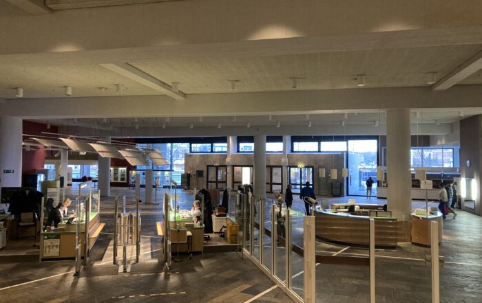 Die geräumige, moderne Lobby der Hochschule Coburg verfügt über große Fenster, die den Bereich mit natürlichem Licht durchfluten. Sie verfügt über einen eleganten Empfangstresen, Glasbarrieren und viele Menschen, die sich dort tummeln. Die dunklen Bodenfliesen bilden einen eleganten Kontrast zu den hellen Wänden und schaffen eine einladende Atmosphäre.