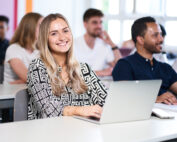 Eine Frau mit langen blonden Haaren lächelt in einem hellen, gut beleuchteten Hörsaal der Hochschule Coburg in die Kamera. Vor ihr liegt ihr aufgeklappter Laptop. Im Hintergrund sind mehrere andere Studierende in eine lebhafte Vorlesung oder Diskussion vertieft.