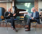 Zwei Männer sitzen auf Stühlen und diskutieren mit Mikrofonen in der Hochschule Coburg. Im Hintergrund sind ein Flügel und Notenständer zu sehen. Einer gestikuliert, während er in dem gemauerten Raum spricht, in dem auch eine Frau sitzt.