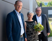 Drei formell gekleidete Menschen stehen im Freien vor dem modernen Gebäude der Hochschule Coburg. Die Frau in der Mitte hält einen Strauß gelber und weißer Blumen in der Hand, im Hintergrund ist üppiges Grün zu sehen.