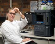 Eine Frau im weißen Laborkittel, Vertreterin der Hochschule Coburg, sitzt an einem Arbeitsplatz in einem Labor. Neben ihr stehen Analysegeräte, sie hält ein kleines Fläschchen und blickt in die Kamera. Auf der Maschine sind Flaschen mit Flüssigkeit zu sehen.