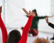 Eine Gruppe von Menschen praktiziert Yoga in Innenräumen der Hochschule Coburg. Eine Person im Vordergrund trägt ein rotes Hemd und hat die Arme erhoben, während andere im Hintergrund verschwommen im Schneidersitz mit ausgestreckten Armen sitzen. Die Umgebung ist hell und natürlich.