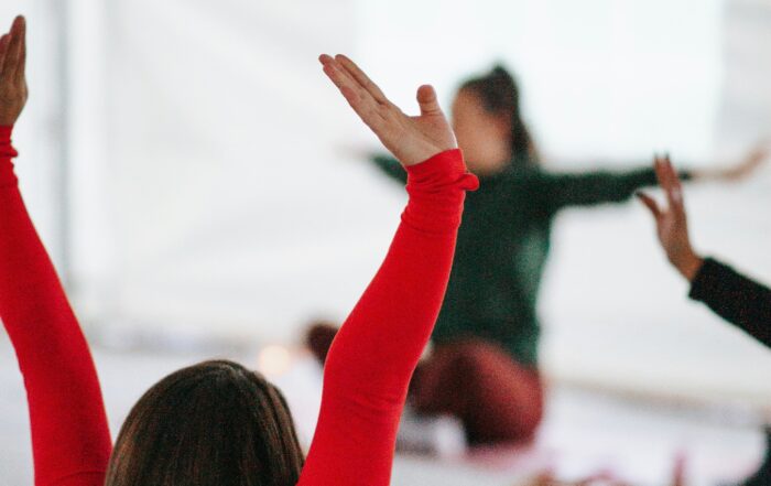 Eine Gruppe von Menschen praktiziert Yoga in Innenräumen der Hochschule Coburg. Eine Person im Vordergrund trägt ein rotes Hemd und hat die Arme erhoben, während andere im Hintergrund verschwommen im Schneidersitz mit ausgestreckten Armen sitzen. Die Umgebung ist hell und natürlich.