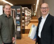 Zwei Männer lächeln in einer modernen Bibliothek der Hochschule Coburg. Einer hält Ordner in der Hand und trägt Brille und Anzug, während der andere ein legeres Hemd trägt. Sie stehen in einem mit Bücherregalen gesäumten Korridor und verkörpern die Mischung aus Professionalität und entspanntem Lernen an dieser renommierten Institution.