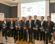 Eine Gruppe von Männern in formellen Anzügen steht in einer Reihe und hält Dokumente in den Händen. Sie befinden sich in einem Konferenzraum mit einem Präsentationsbildschirm, auf dem neben den Logos verschiedener Unternehmen und der Hochschule Coburg der Titel „Shuttle-Modellregion Oberfranken“ zu sehen ist.