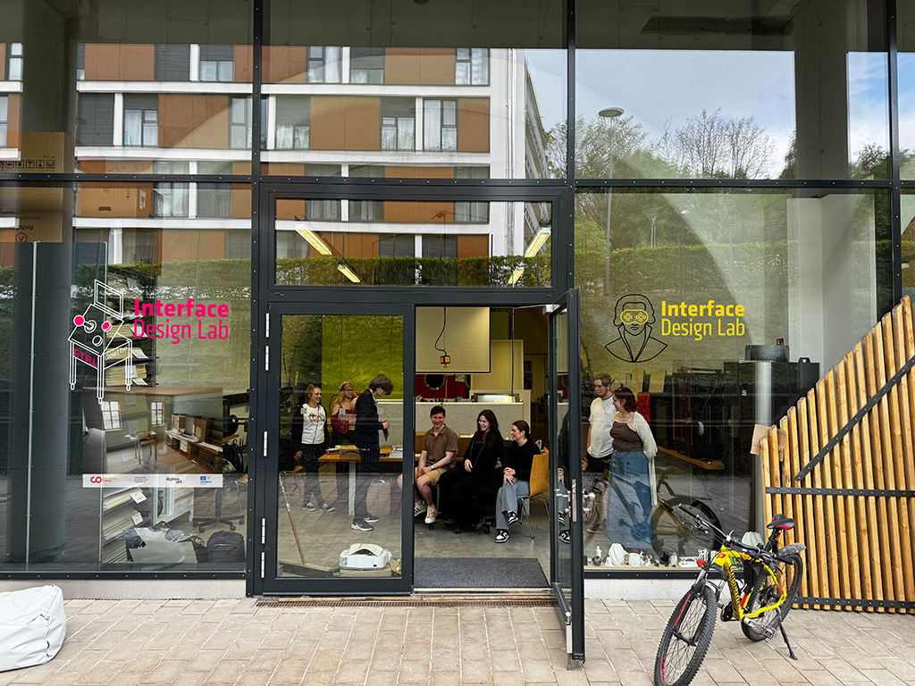 A group of people are gathered inside a building with large glass doors labeled "Interface Design Lab" at Hochschule Coburg. Outside, a bicycle is parked on the pavement, while reflections of trees and nearby campus buildings appear on the glass.