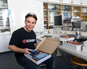 Eine Person in einem Labor der Hochschule Coburg lächelt, während sie einen offenen Karton mit elektronischen Bauteilen in der Hand hält. Der Raum ist mit Regalen, einem Whiteboard und verschiedenen elektronischen Geräten und Werkzeugen auf einem Schreibtisch ausgestattet.