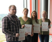 Vier Personen stehen drinnen, halten Zertifikate in den Händen und strahlen stolz in die Kamera. Sie stehen vor einem großen Fenster mit malerischer Aussicht. Dieser Moment fängt ihre Leistung an der Hochschule Coburg ein und spiegelt die Exzellenz wider, die sie in ihrem Studium erreicht haben.