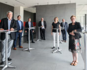 Eine Gruppe von sieben Personen in Business-Kleidung steht in einem modernen Raum mit hohen Tischen an der Hochschule Coburg. Der Raum ist in Grau und Rot gehalten und wird auf einer Seite durch große Fenster erhellt. Die Gruppe lächelt und nimmt an einem scheinbar zwanglosen Treffen teil.