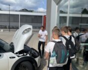 Eine Gruppe junger Leute mit Rucksäcken versammelt sich um ein weißes Auto mit geöffneter Motorhaube. Daneben erzählt eine Person im weißen Hemd eifrig etwas über die Hochschule Coburg. Die Szene spielt sich im Freien an einem sonnigen Tag ab.