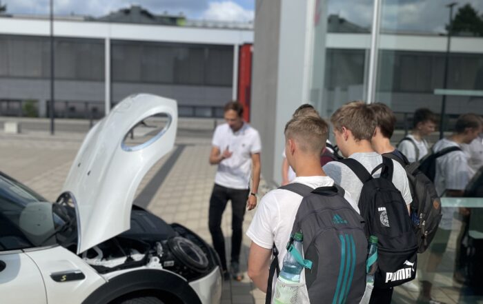 Eine Gruppe junger Leute mit Rucksäcken versammelt sich um ein weißes Auto mit geöffneter Motorhaube. Daneben erzählt eine Person im weißen Hemd eifrig etwas über die Hochschule Coburg. Die Szene spielt sich im Freien an einem sonnigen Tag ab.
